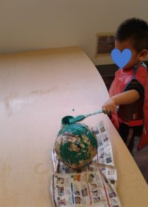 Child paints a big paper mache ball with green paint and a brush.