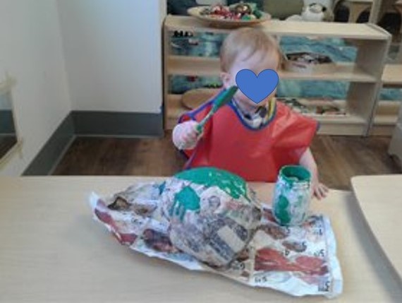 Child paints a big paper mache ball with green paint and a brush.